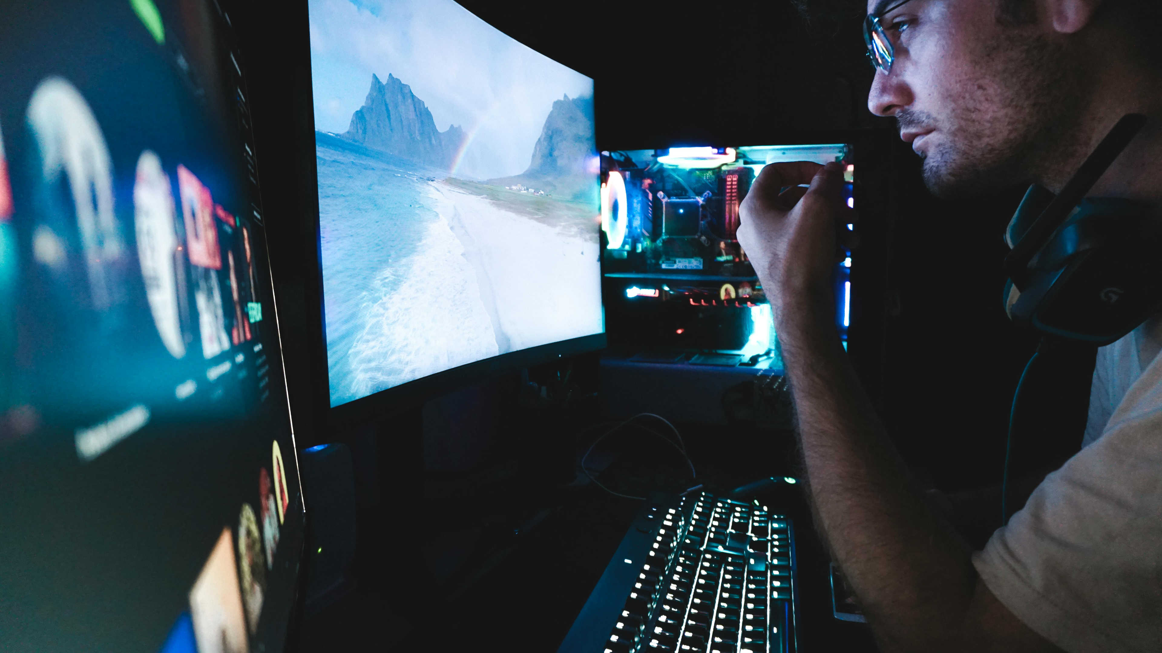 A man looking at his two screens with glasses in a dark room and his custom PC glows in the background