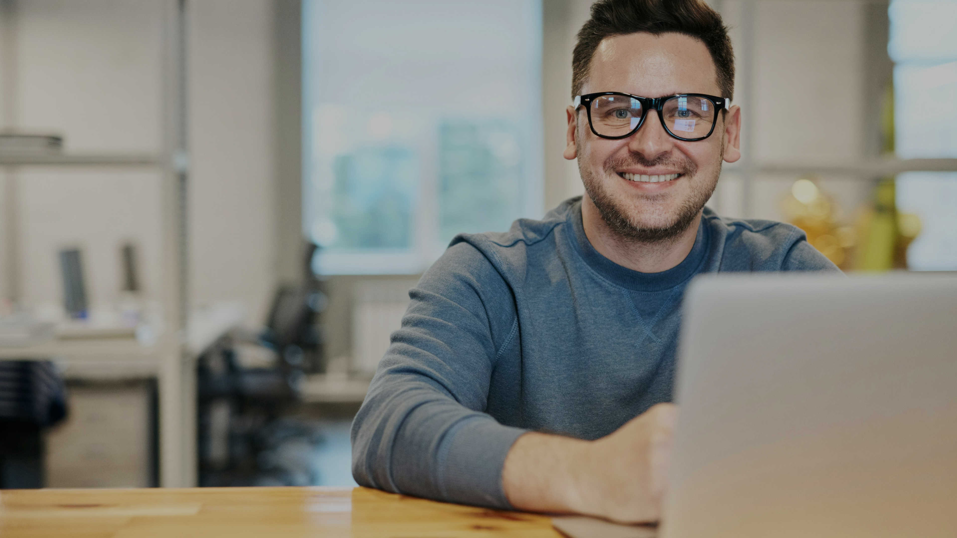 A man with glasses sitting at a desk in front of a laptop smiling at the camera