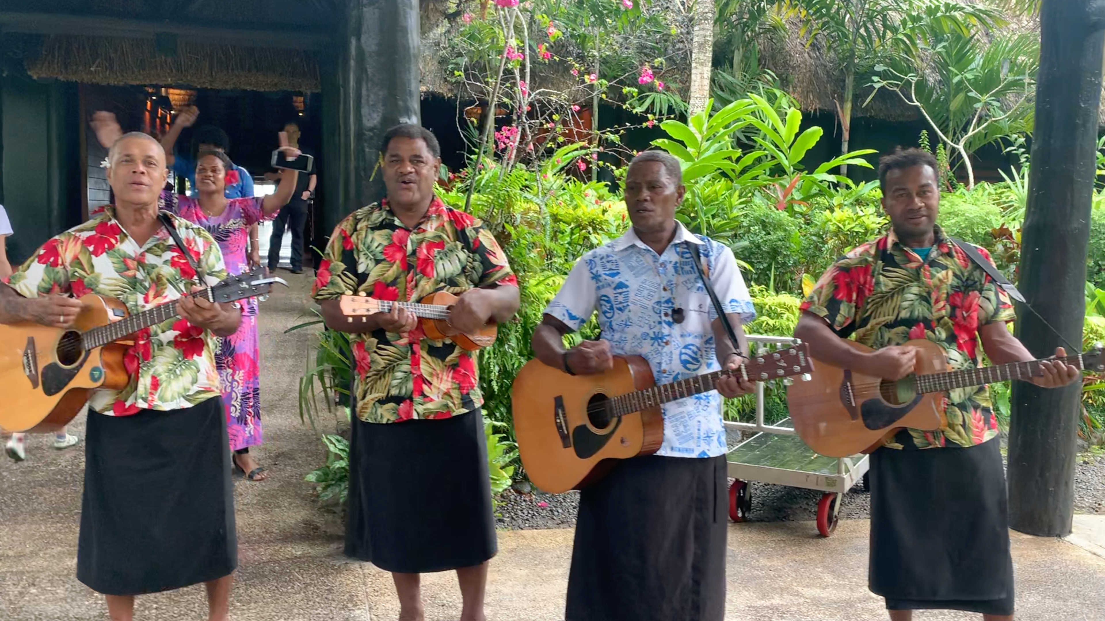 Four members of the resort's own Bula Band with guitars and a ukelele and colorful shirts singing and playing music as women wave goodbye behind them.