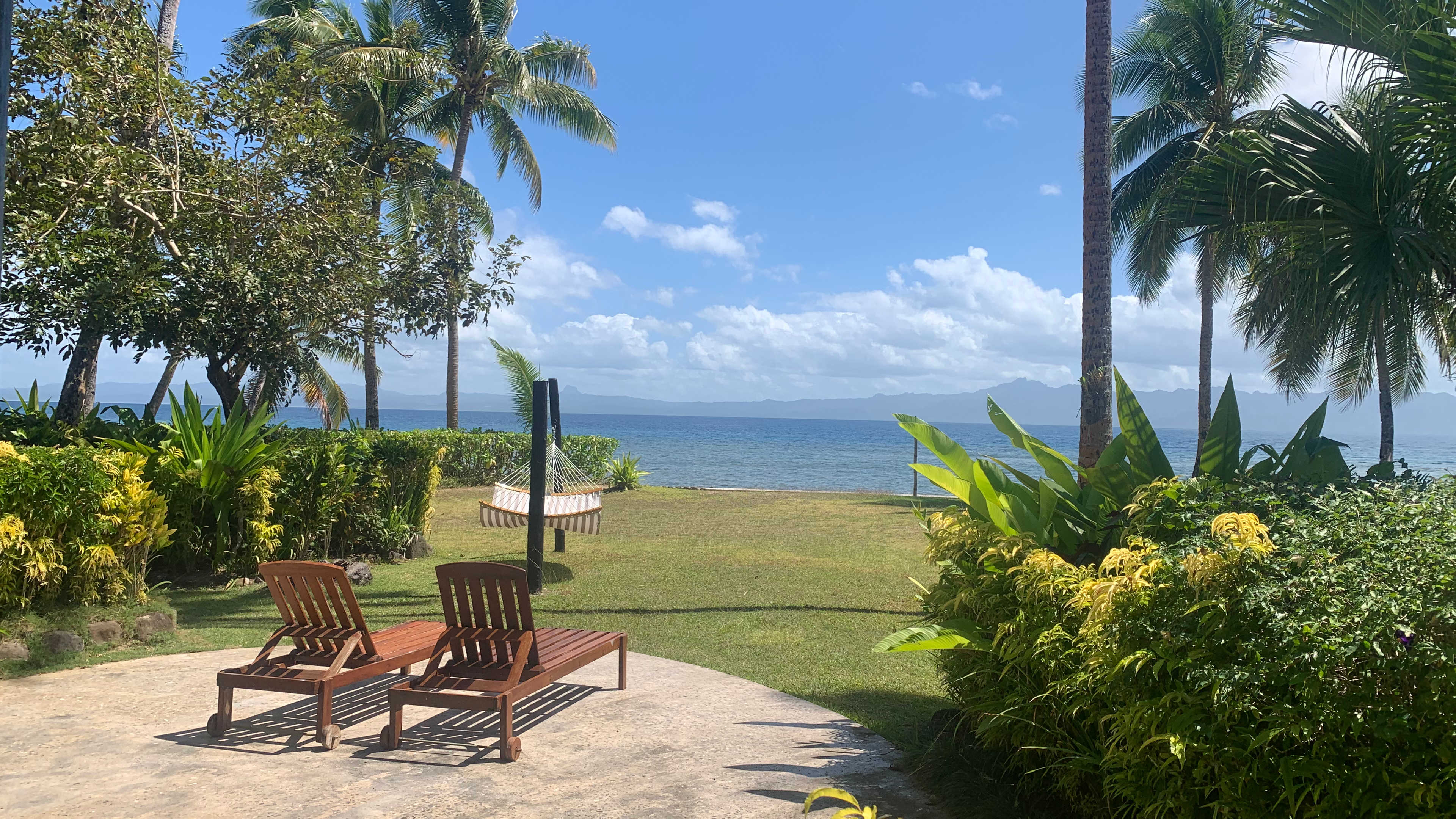 The view from our bure including palm trees, tropical flowers and greenery, manicured lawn, two chaise lounges, a hammock and Savusavu Bay in the background on a beautiful day