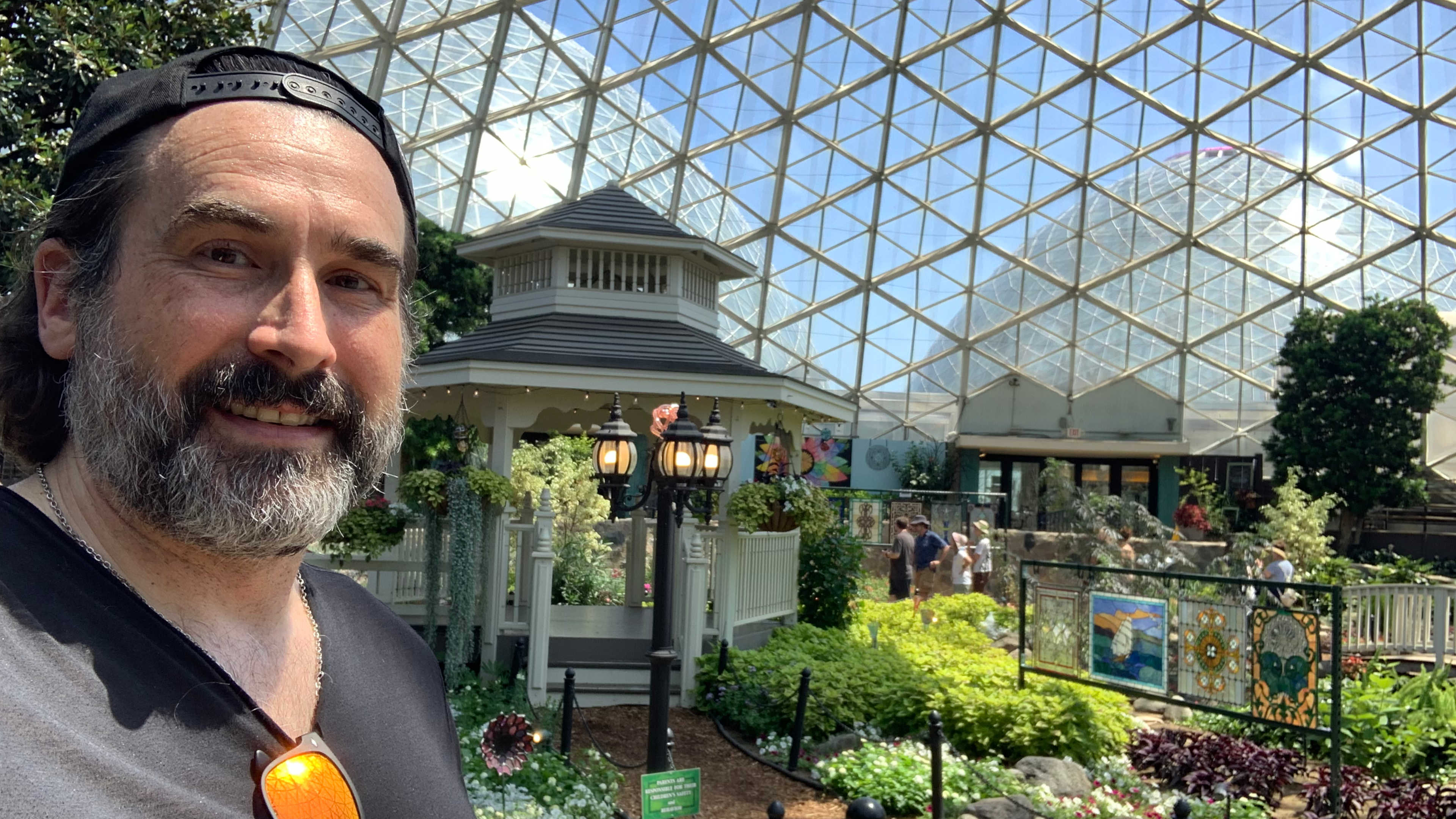Self-portrait inside the glass-domed conservatory with a gazebo, garden beds and visitors in the background.