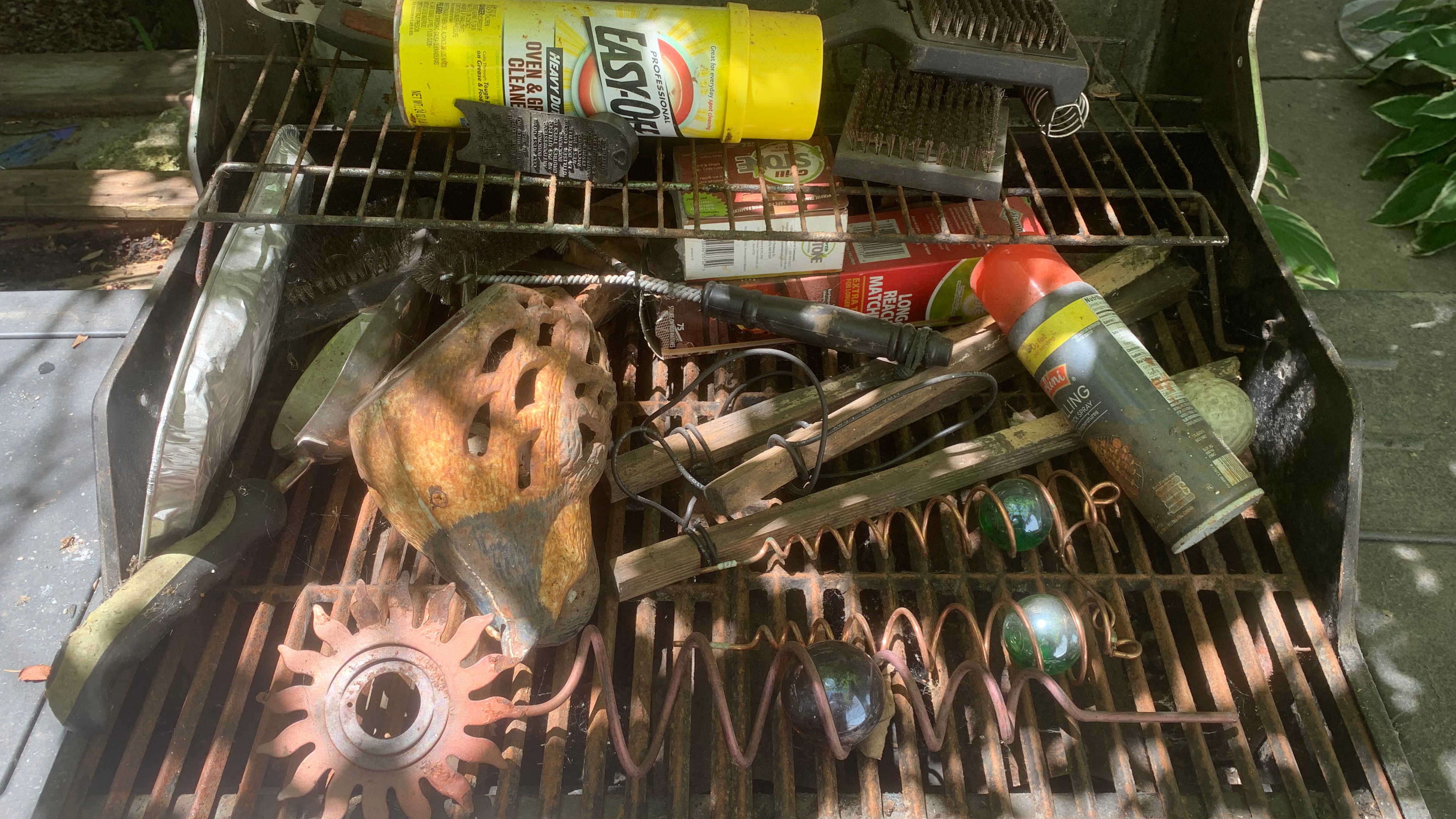 Outdoor grill filled with rusted grates, cleaning supplies and stored garden objects before restoration.